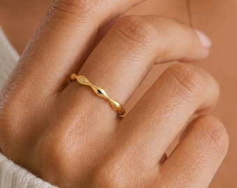 May include: A thin gold ring featuring a repeating series of small, polished, almond-shaped metal segments connected in a continuous band. The ring has a bright, reflective finish and a delicate, uniform structure that encircles the finger.