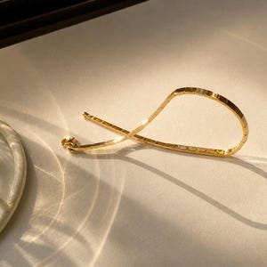 Pu&ograve; includere: Un bracciale color oro composto da una serie di maglie rettangolari piatte collegate tra loro. La catena presenta una finitura lucida che riflette la luce, creando una forma curva e flessibile con un meccanismo di chiusura visibile.