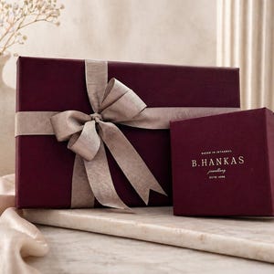 Pu&ograve; includere: Una scatola regalo rettangolare color bordeaux avvolta da un nastro beige annodato a fiocco, posizionata accanto a una scatola quadrata pi&ugrave; piccola color bordeaux con il testo dorato B.HANKAS jewellery ESTD 1998. Le scatole hanno una finitura opaca.