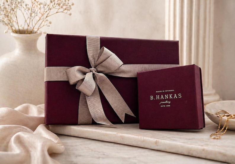 Pu&ograve; includere: Una scatola regalo rettangolare color bordeaux avvolta da un nastro beige annodato a fiocco, posizionata accanto a una scatola quadrata pi&ugrave; piccola color bordeaux con il testo dorato B.HANKAS jewellery ESTD 1998. Le scatole hanno una finitura opaca.