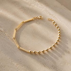 Puede incluir: Una pulsera de tono dorado con un diseño de cadena doble. Una mitad de la pulsera consta de eslabones de barra rectangulares y lisos, mientras que la otra mitad está compuesta por pequeñas cuentas esféricas redondas y un cierre de resorte.
