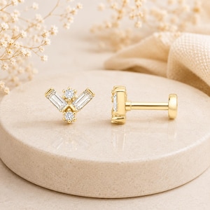 May include: A pair of gold-colored stud earrings with a geometric design. Each earring features a cluster of baguette-cut and round-cut clear stones. One earring is shown from the side, displaying the post and backing.