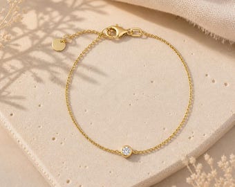 Può includere: Un delicato bracciale a catena dorata con una singola pietra preziosa trasparente rotonda incastonata a lunetta al centro. Il bracciale include una sottile catena a maglie, una chiusura a moschettone sicura e una piccola targhetta circolare.