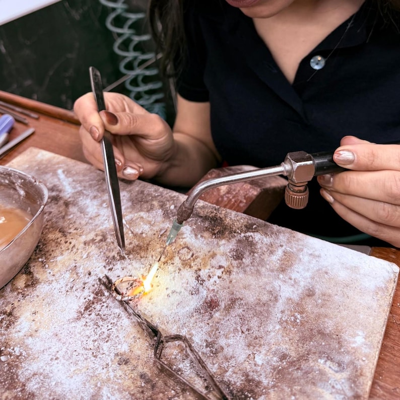 Pu&ograve; includere: Un anello in metallo viene saldato su una superficie da lavoro ruvida e usurata utilizzando un cannello manuale che emette una piccola fiamma luminosa. Una pinzetta metallica lunga e sottile tiene fermo l'anello durante il riscaldamento.