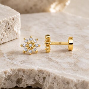 Peut inclure: Une boucle d'oreille à tige en or en forme de fleur avec huit petites pierres claires disposées en cercle. La tige est visible, portant l'inscription gravée 14K, et comprend un embout circulaire plat fixé à l'extrémité de la tige.