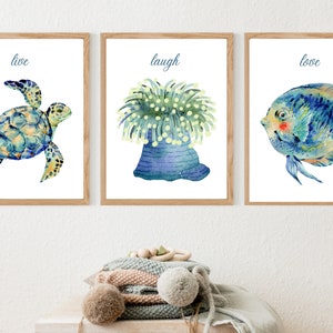 Puede incluir: Tres impresiones de acuarela con una tortuga marina azul y verde, una anémona azul y verde y un pez azul y verde. Las impresiones están enmarcadas en madera natural y tienen las palabras "vivir", "reír" y "amar" impresas debajo de las imágenes.