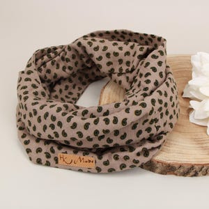 Könnte beinhalten: Ein beigefarbener Loop-Schal mit einem Paisley-Muster in Dunkelgrün. Der Schal ist aus weichem Stoff gefertigt und hat ein kleines Lederetikett mit der Aufschrift "H U Made". Eine große weiße Blume liegt neben dem Schal auf einer Holzscheibe.