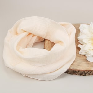 Könnte beinhalten: Ein pfirsichfarbener Rundschal aus leichtem, weichem Stoff wird auf einer Holzscheibe neben einer großen, weißen Blume präsentiert. Der Schal ist kreisförmig drapiert. Die Blume hat mehrere Blütenblätter.