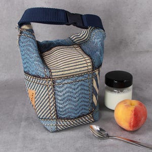 Könnte beinhalten: Eine Lunchtasche aus Patchwork-Denim mit blauem Griff und Schnallenverschluss. Daneben ein Glas mit schwarzem Deckel, ein Pfirsich und ein Löffel. Die Tasche hat verschiedene Denim-Muster.