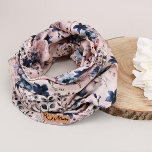 Könnte beinhalten: Ein rosa-blauer Blumen-Loop-Schal. Der Schal hat ein sich wiederholendes Muster aus blauen Blumen und grünen Blättern auf rosa Grund. Ein kleines braunes Etikett mit Text ist angebracht. Der Schal liegt auf einer Holzscheibe.