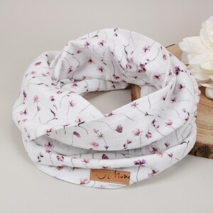 Könnte beinhalten: Ein weißer Loop-Schal mit zartem Blumenmuster in Rosa- und Lilatönen. Der Schal ist aus weichem Stoff gefertigt und hat ein kleines braunes Lederetikett. Eine große weiße Blume liegt daneben.