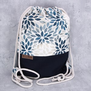 Könnte beinhalten: Ein Turnbeutel mit floralem Muster in Blau- und Beigetönen im oberen Bereich und einem marineblauen Boden. Die Tasche hat weiße Kordelträger und ein kleines braunes Lederetikett. Ideal für Sport oder Freizeit.