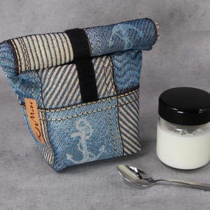Könnte beinhalten: Eine Lunchtasche aus Jeansstoff mit schwarzem Riemen, ein kleines Glas mit schwarzem Deckel und ein silberner Löffel. Die Tasche hat ein Patchwork-Design mit Anker- und Streifenmustern. Das Glas ist mit einer weißen Flüssigkeit gefüllt.