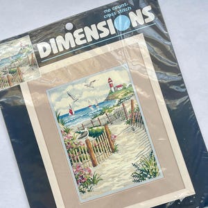 May include: A Dimensions no count cross stitch kit titled Lovely Seascape featuring a coastal scene with a lighthouse, sailboats, seagulls, and a wooden fence along a sandy path. The package indicates a finished frame size of 14 by 18 inches.