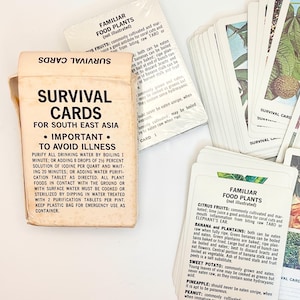 May include: A deck of Survival Cards for South East Asia in a white paper box. The box features black text with instructions for water purification. The cards display colorful illustrations of various plants and fruits with descriptive text.