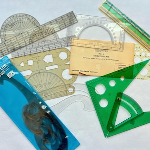 May include: A collection of drafting tools including a Staedtler Mars 571 40 WP French curve in blue packaging, a green triangle with a protractor, a RapiDesign No. 40 circle template, various clear plastic protractors, and a long green ruler.