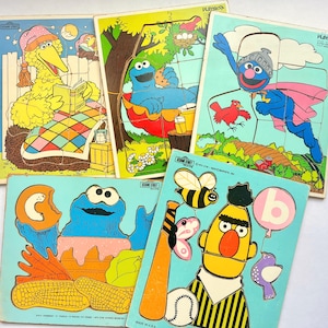 May include: Five vintage Sesame Street wooden inlay puzzles featuring Big Bird reading in bed, Cookie Monster eating a cookie in a hammock, Super Grover flying, Cookie Monster with food, and Bert with a baseball bat, ball, bee, and balloon.