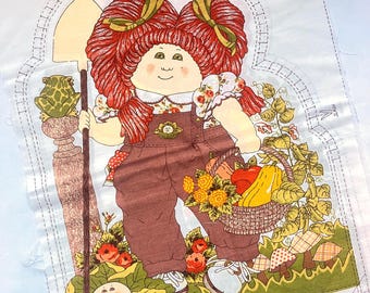 May include: A fabric panel with a Cabbage Patch Kid design. The illustration shows a character with red pigtails, brown overalls, and a shovel, with a basket of fruit and vegetables. Includes instructions and the text "LEAVE OPEN FOR STUFFING".