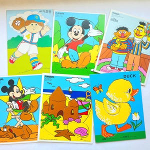 May include: A collection of vintage Playskool jigsaw puzzles featuring cartoon characters. Puzzles include Mickey Mouse, Ernie and Bert, a baseball player, and a yellow duck. Each puzzle has a colorful design and is made of cardboard.