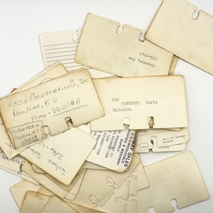 May include: A collection of vintage Rolodex cards in various sizes and conditions. Some cards have handwritten information, including names, phone numbers, and company names. The cards are beige and show signs of age.