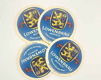 May include: Four round Löwenbräu beer coasters. Each coaster features a blue background with a yellow lion emblem and the words "Löwenbräu Munich Imported German Beer". The coasters are made in Germany.