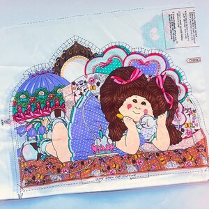 May include: A fabric panel featuring a colorful illustration of a doll with brown hair and pink bows, lying on a patterned bed. The doll is holding a spoon and a small container. The panel includes instructions and the word "Cabbage".