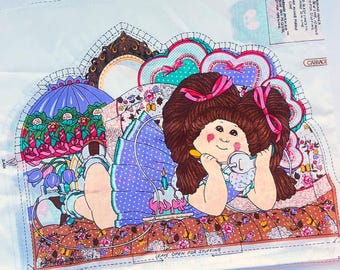 May include: A fabric panel with a colourful illustration of a doll with brown hair and pink bows, lying on a patterned bed. The doll is holding a spoon and a small container. The panel includes instructions and the word "Cabbage".