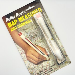 May include: A Roller Route map measurer and compass in its original packaging. The packaging features the text "Roller Route MAP MEASURER AND COMPASS". The map measurer is white with red accents and a black scale. The packaging is on a white background.