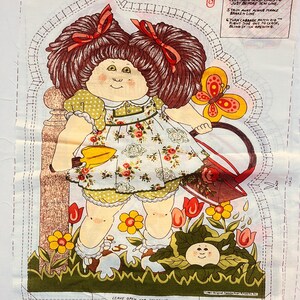 May include: A vintage Cabbage Patch Kids fabric panel featuring a doll design. The doll has brown hair with red bows, a floral dress, and is surrounded by flowers and a butterfly. Instructions are printed on the fabric.