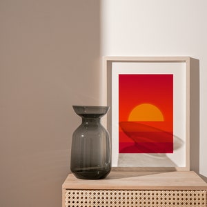 May include: A gray glass vase sits on a light wood nightstand with a woven rattan drawer. A framed print of a sunset with orange and red hues hangs above the vase.