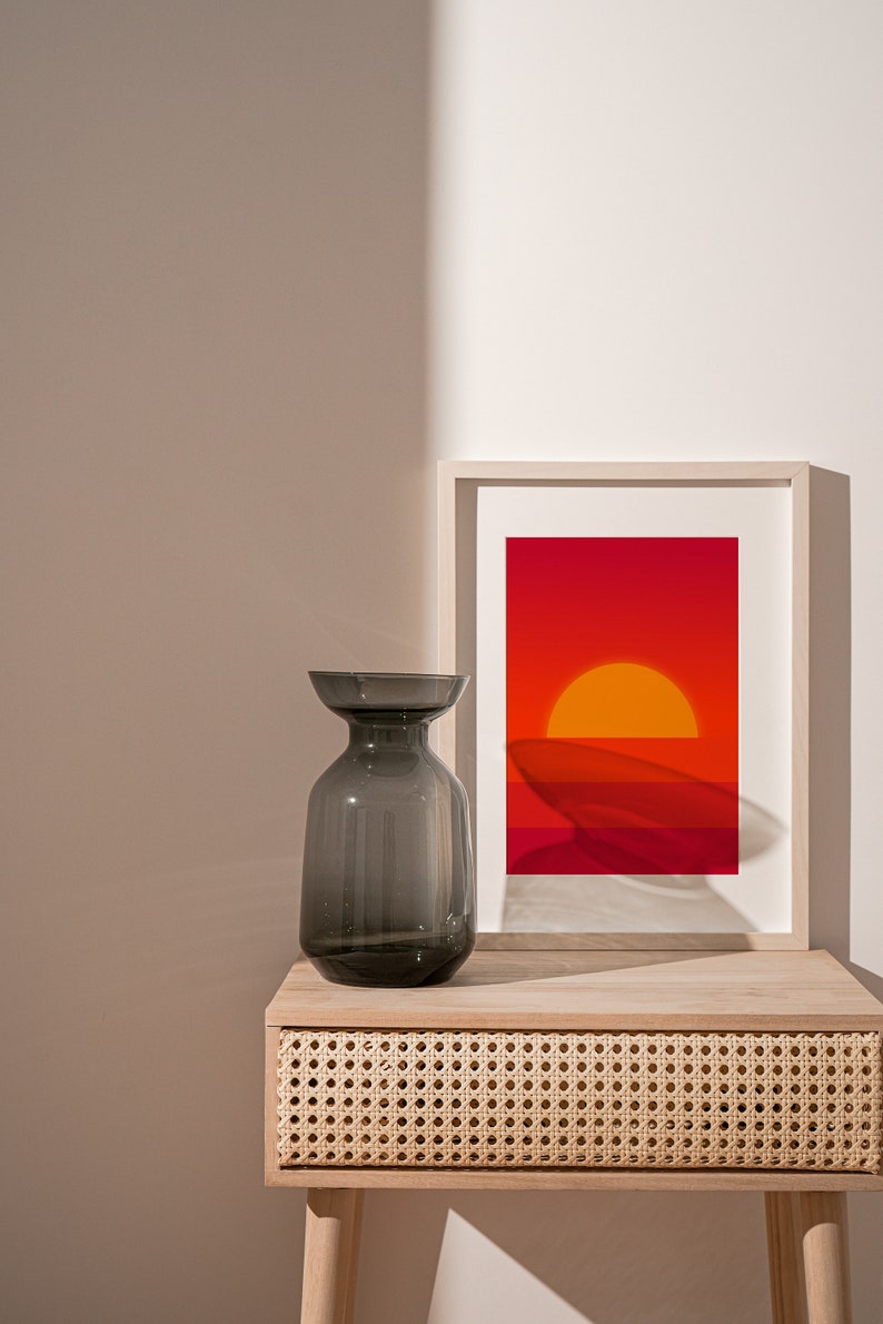 May include: A gray glass vase sits on a light wood nightstand with a woven rattan drawer. A framed print of a sunset with orange and red hues hangs above the vase.