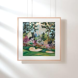May include: A framed painting of a golf course scene. The artwork depicts a green fairway with two sand traps, surrounded by colorful flowers and trees. The painting is in a light wooden frame, hanging on a white wall.