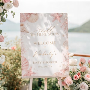 May include: A rectangular white welcome sign for a baby shower featuring a soft pink seashell and starfish border. The text reads: Shell BE HERE SOON, WELCOME TO, Kimberly's, BABY SHOWER, 09.20.YY. The sign is displayed on a wooden easel stand.