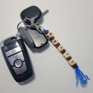 May include: A keychain with a blue tassel and wooden beads spelling out the word "BRAVE". The keychain is attached to a set of keys, including a black car key fob and a silver key.