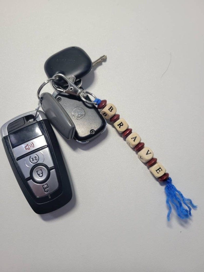 May include: A keychain with a blue tassel and wooden beads spelling out the word "BRAVE". The keychain is attached to a set of keys, including a black car key fob and a silver key.