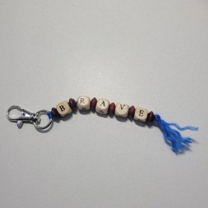 May include: A wooden keychain with the word "BRAVE" spelled out in black letters on individual beads. The beads are strung on a blue cord with a silver metal clasp.