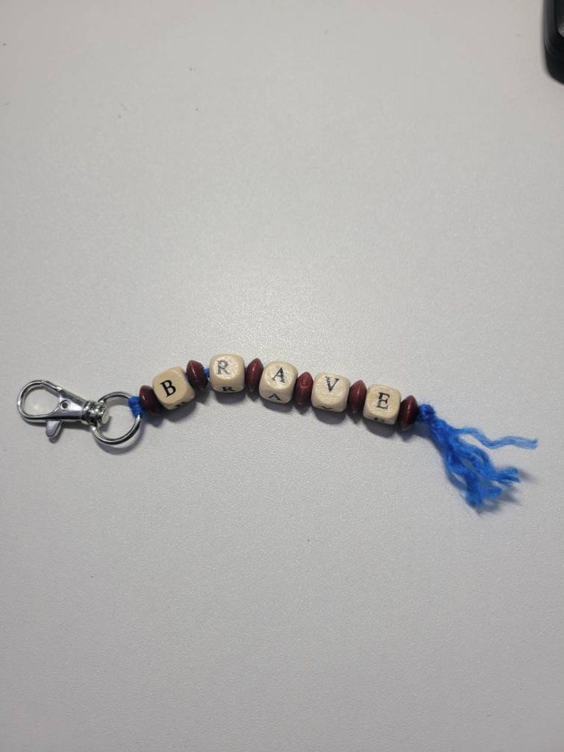May include: A wooden keychain with the word "BRAVE" spelled out in black letters on individual beads. The beads are strung on a blue cord with a silver metal clasp.