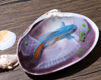 Puede incluir: Una figurita de pez azul y naranja está encapsulada en resina dentro de una concha de mar. Puntos de resina verde están dispersos alrededor del pez.