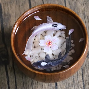 May include: A wooden bowl filled with water, white pebbles, and two resin fish. One fish is white with black spots, and the other is black with white spots. A pink flower is in the center of the bowl.