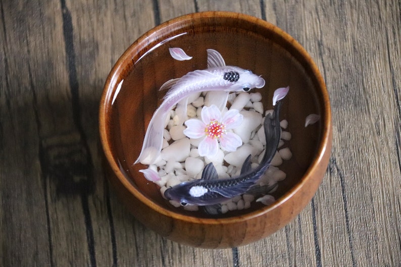 May include: A wooden bowl filled with water, white pebbles, and two resin fish. One fish is white with black spots, and the other is black with white spots. A pink flower is in the center of the bowl.