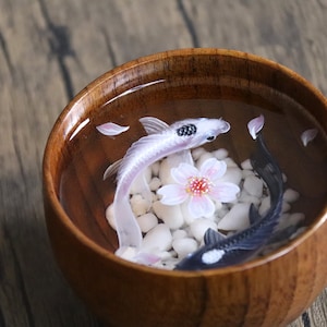 May include: A wooden bowl filled with water, white pebbles, and two koi fish. One fish is white with black markings, and the other is black with white markings. A pink cherry blossom is floating in the water.