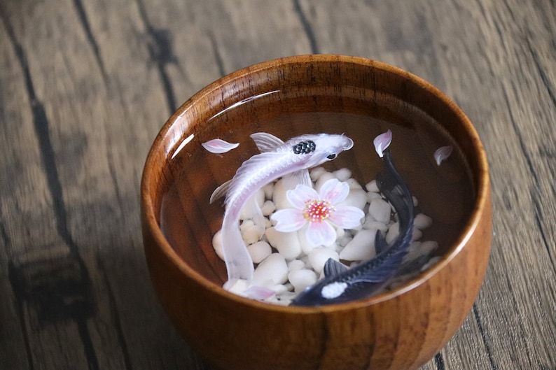 May include: A wooden bowl filled with water, white pebbles, and two koi fish. One fish is white with black markings, and the other is black with white markings. A pink cherry blossom is floating in the water.