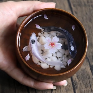 May include: A wooden bowl filled with water, white pebbles, and pink cherry blossoms. Two koi fish, one black and one white, swim in the bowl.