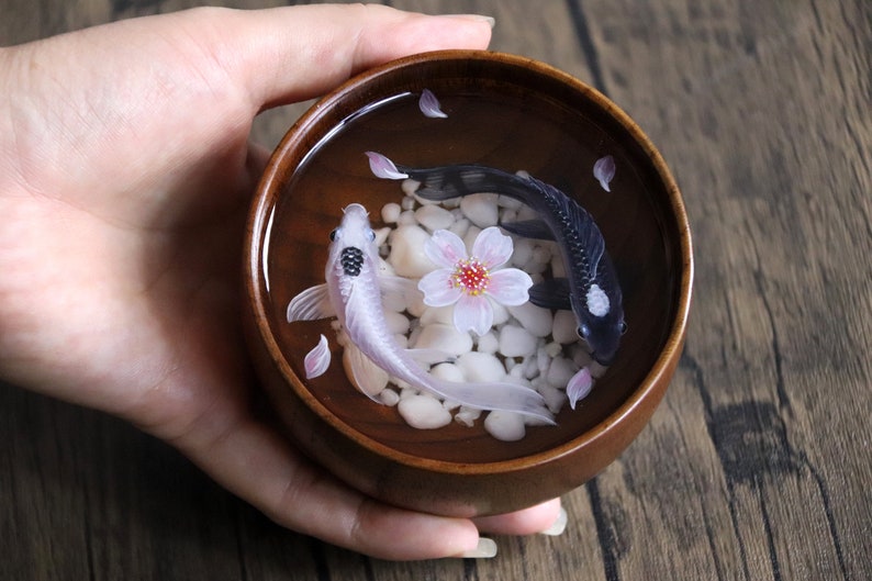 May include: A wooden bowl filled with water, white pebbles, and pink cherry blossoms. Two koi fish, one black and one white, swim in the bowl.