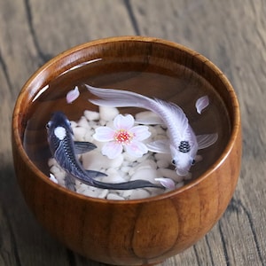 May include: A wooden bowl filled with clear resin, containing two koi fish, one black and one white, with a pink cherry blossom in the center. White pebbles and pink petals are scattered around the fish.