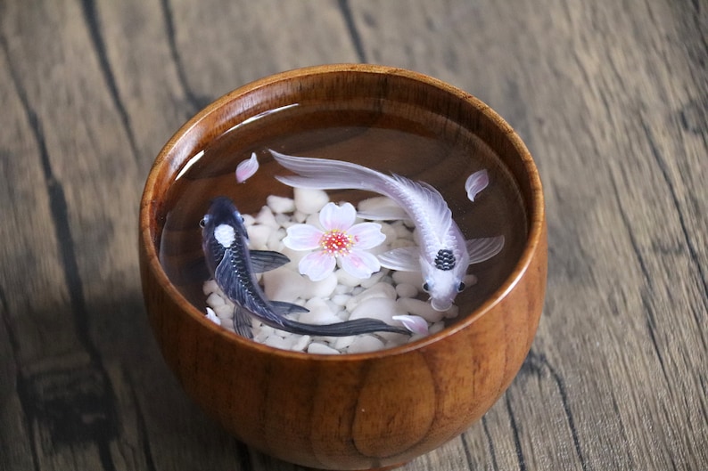 May include: A wooden bowl filled with clear resin, containing two koi fish, one black and one white, with a pink cherry blossom in the center. White pebbles and pink petals are scattered around the fish.