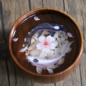 May include: Two resin koi fish, one black and one white, swim in a wooden bowl filled with water and white stones. A pink cherry blossom sits in the center of the bowl.