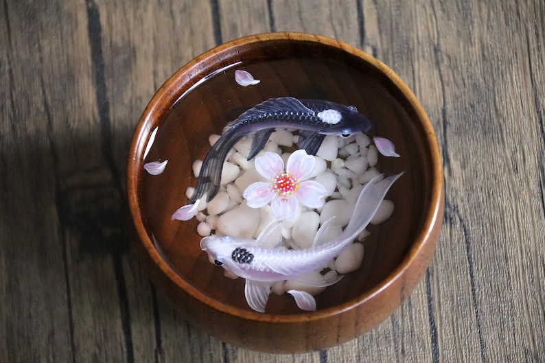 May include: Two resin koi fish, one black and one white, swim in a wooden bowl filled with water and white stones. A pink cherry blossom sits in the center of the bowl.