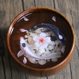 May include: A wooden bowl filled with water, white stones, and two koi fish. One fish is black with white spots, and the other is white with black spots. A pink cherry blossom is in the center of the bowl.