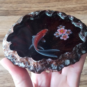 May include: A red and white goldfish painted on a dark brown agate slice with pink flowers.